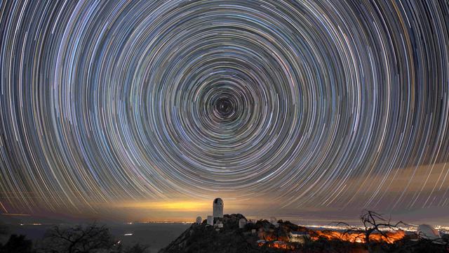 Circles of light on the night sky. A telescope dome atop a mountain is below the center of the circle.